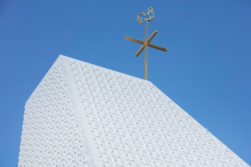 Fassadendetail der Kirche St. Michael in Poing mit weißer, geometrisch strukturierter Oberfläche aus dreidimensionalen, pyramidenartigen Fassadenkacheln vor klarem, blauem Himmel