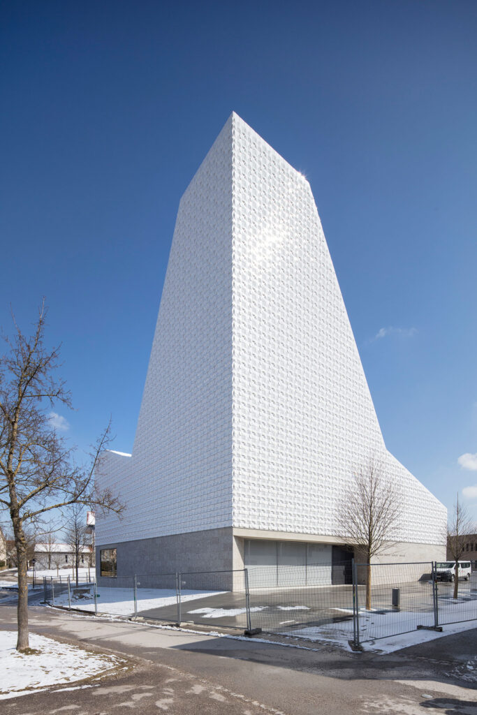 Fassadendetail der Kirche St. Michael in Poing mit weißer, geometrisch strukturierter Oberfläche aus dreidimensionalen, pyramidenartigen Fassadenkacheln vor klarem, blauem Himmel