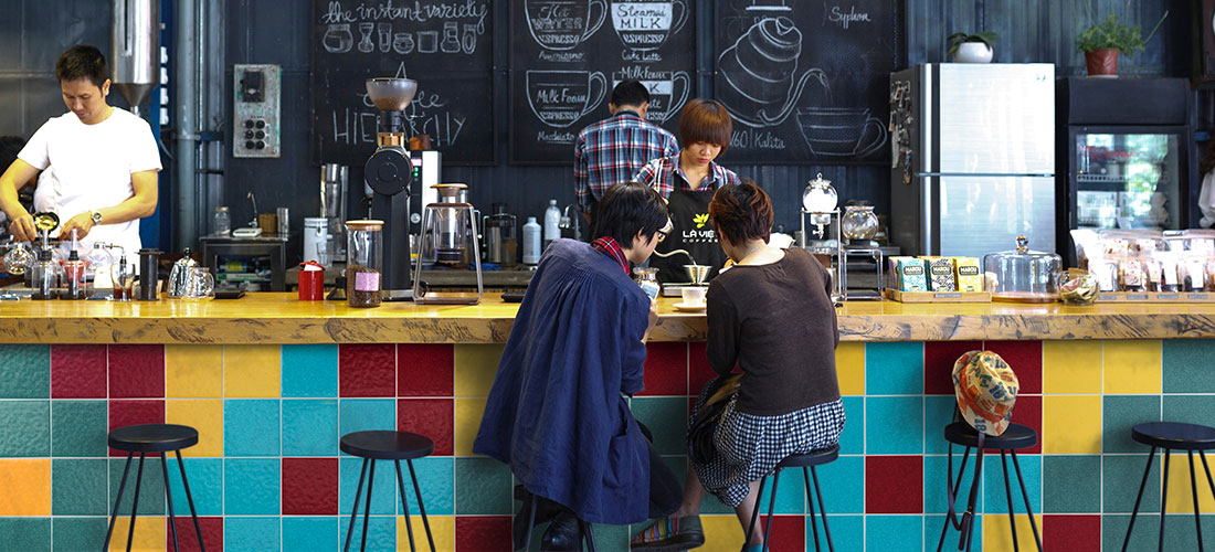 Zwei Personen sitzen an einer bunten Bar in einem Café, im Hintergrund Baristas bereiten Getränke vor, Wandtafel mit handgeschriebenen Kaffeespezialitäten