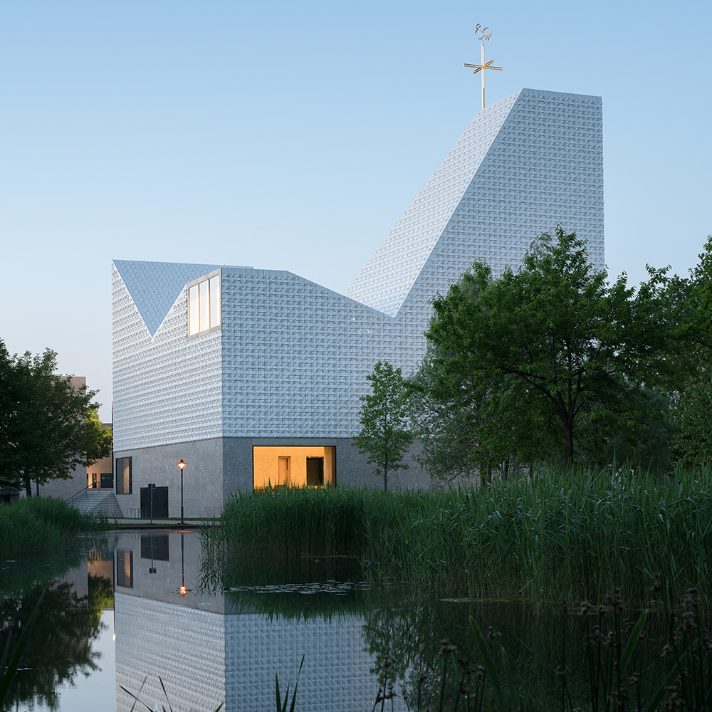 Église St. Michael à Poing avec une surface blanche à structure géométrique composée de carreaux de façade tridimensionnels en forme de pyramide. Vue sur l'étang au crépuscule. 