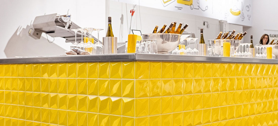 Bar mit gelben, strukturierten Fliesen. Auf der Theke stehen Flaschen, Gläser und Getränkebehälter.