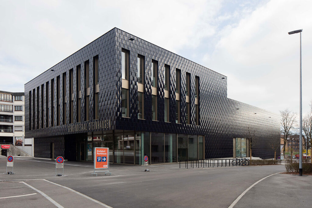 Modernes Gebäude mit schwarzer Fassade und großen Fenstern, Schriftzug 'Gemeindesaal Steinhausen' an der Front