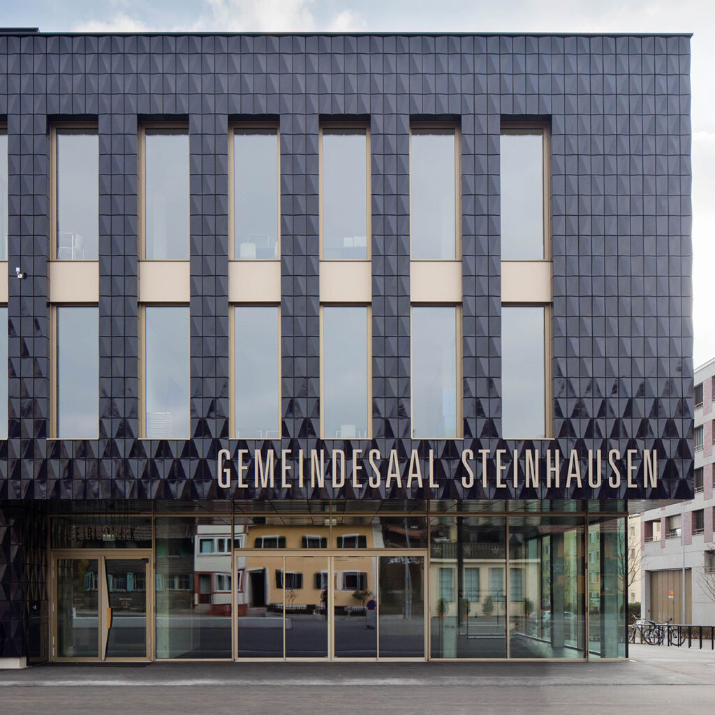 Modernes Gebäude mit schwarzer Fassade und großen Fenstern, Schriftzug 'Gemeindesaal Steinhausen' an der Front