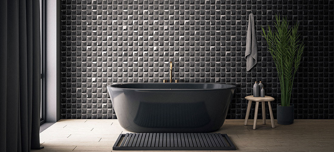 Schwarze freistehende Badewanne vor Wand mit 3D-Fliesen in quadratischem Muster, daneben graues Handtuch, Holzstuhl mit Seifenspendern und hohe Pflanze